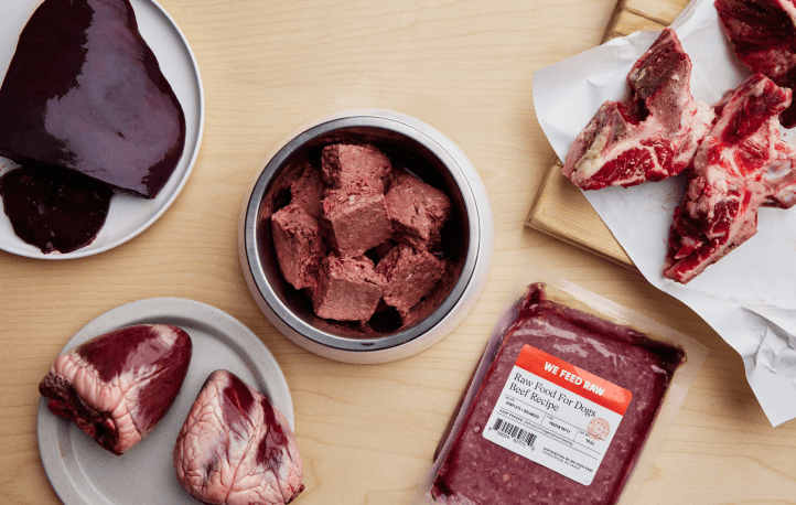 Assortment of raw dog food ingredients including organ meat, raw meat portions, bone-in cuts, and packaged beef blend from We Feed Raw on wooden surface.