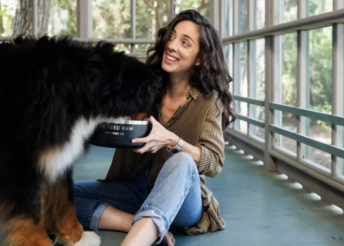 Person sits on a sunlit porch, smiling while feeding a Bernese Mountain Dog from a bowl labeled 'WE FEED RAW'