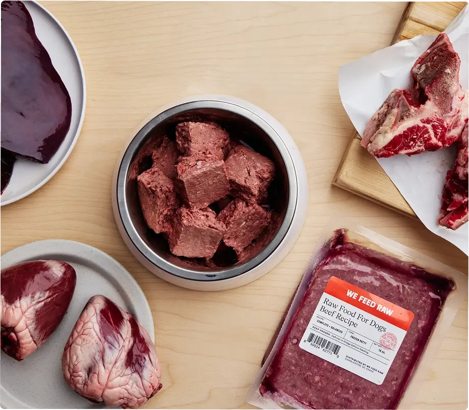 Various cuts of raw meat and prepared raw dog food displayed on a wooden cutting board with butcher knife and kitchen towels. Includes raw muscle meat, organs, and ground meat in a metal container.