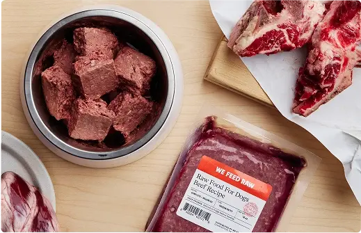 Various cuts of raw meat and prepared raw dog food displayed on a wooden cutting board with butcher knife and kitchen towels. Includes raw muscle meat, organs, and ground meat in a metal container.