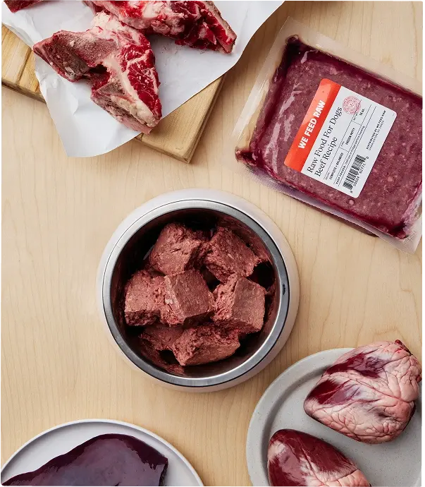 Various cuts of raw meat and prepared raw dog food displayed on a wooden cutting board with butcher knife and kitchen towels. Includes raw muscle meat, organs, and ground meat in a metal container.