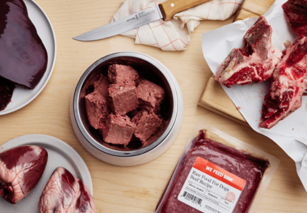 Various cuts of raw meat and prepared raw dog food displayed on a wooden cutting board with butcher knife and kitchen towels. Includes raw muscle meat, organs, and ground meat in a metal container.